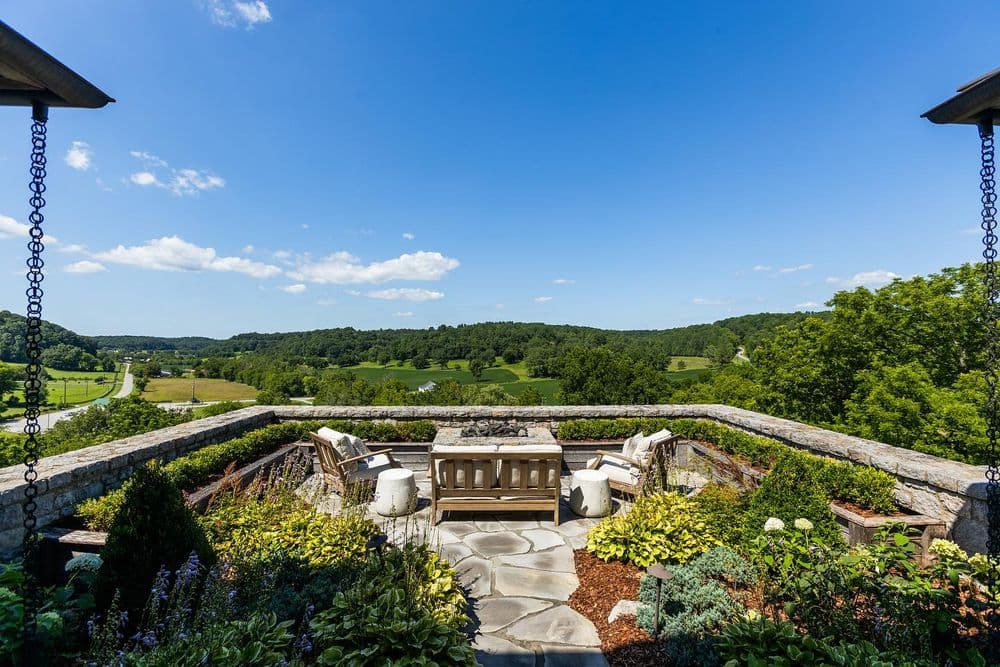 Scenic outdoor terrace with stone table, chairs, and lush greenery overlooking a valley.