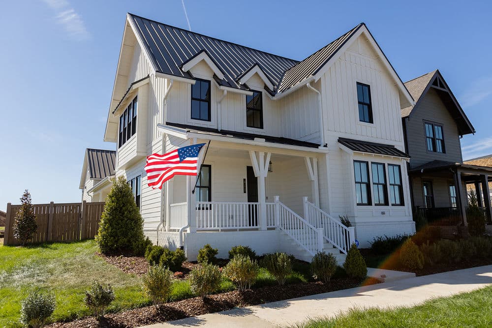 Modern white house with a black metal roof and an American flag in a sunny neighborhood.