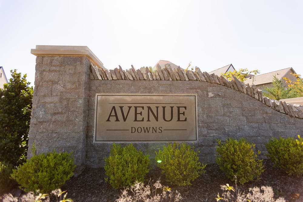Sign for Avenue Downs featuring stone structure and greenery in bright sunlight.