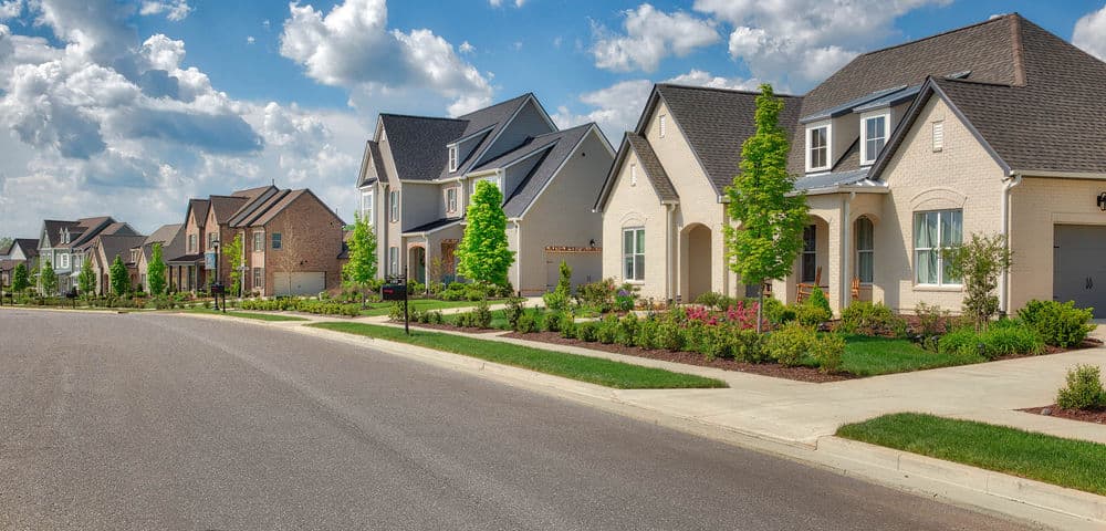 Modern suburban homes line a quiet street under a blue sky with fluffy clouds.