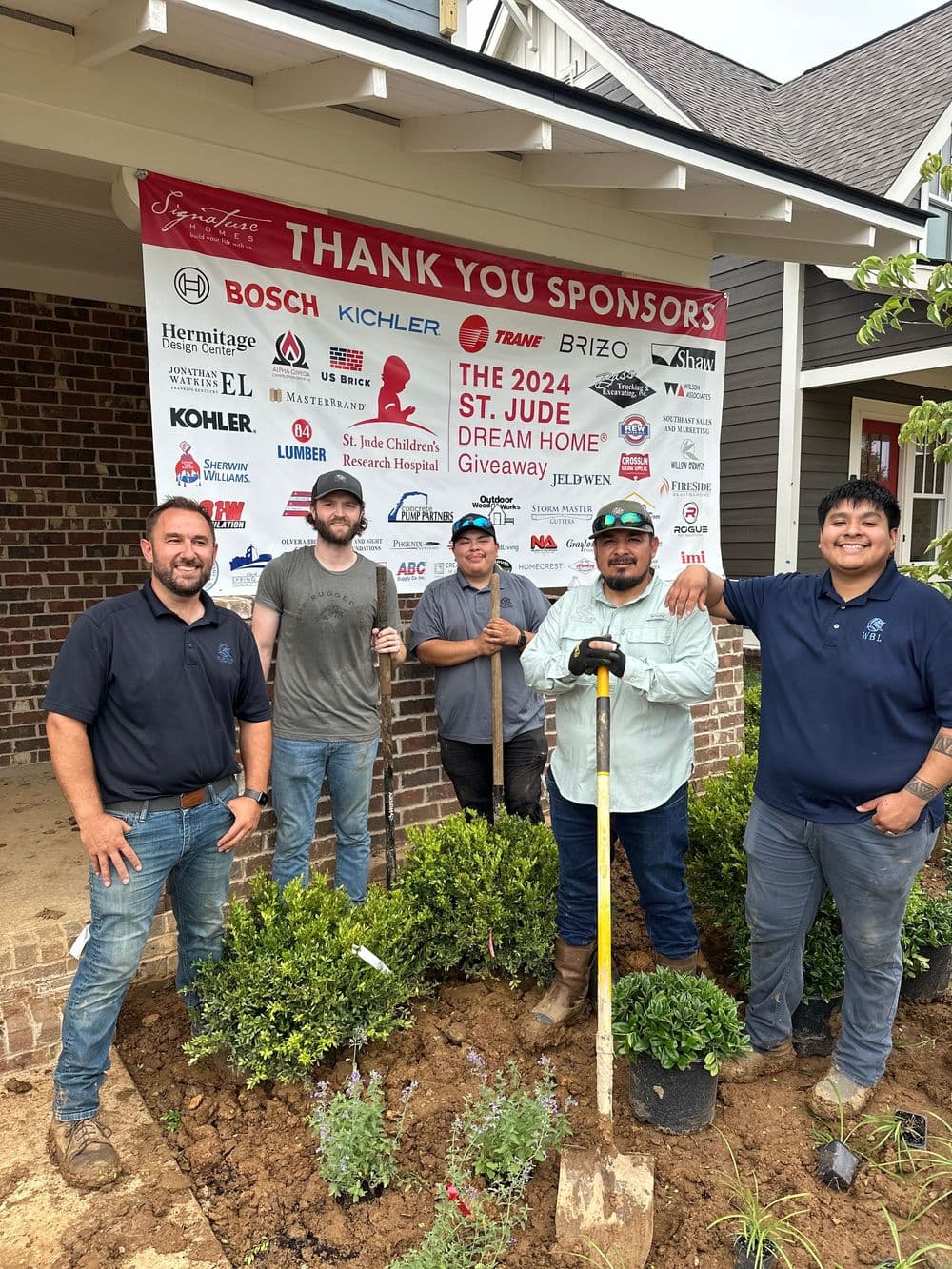 Men posing with gardening tools in front of a St. Jude Dream Home sponsor sign.