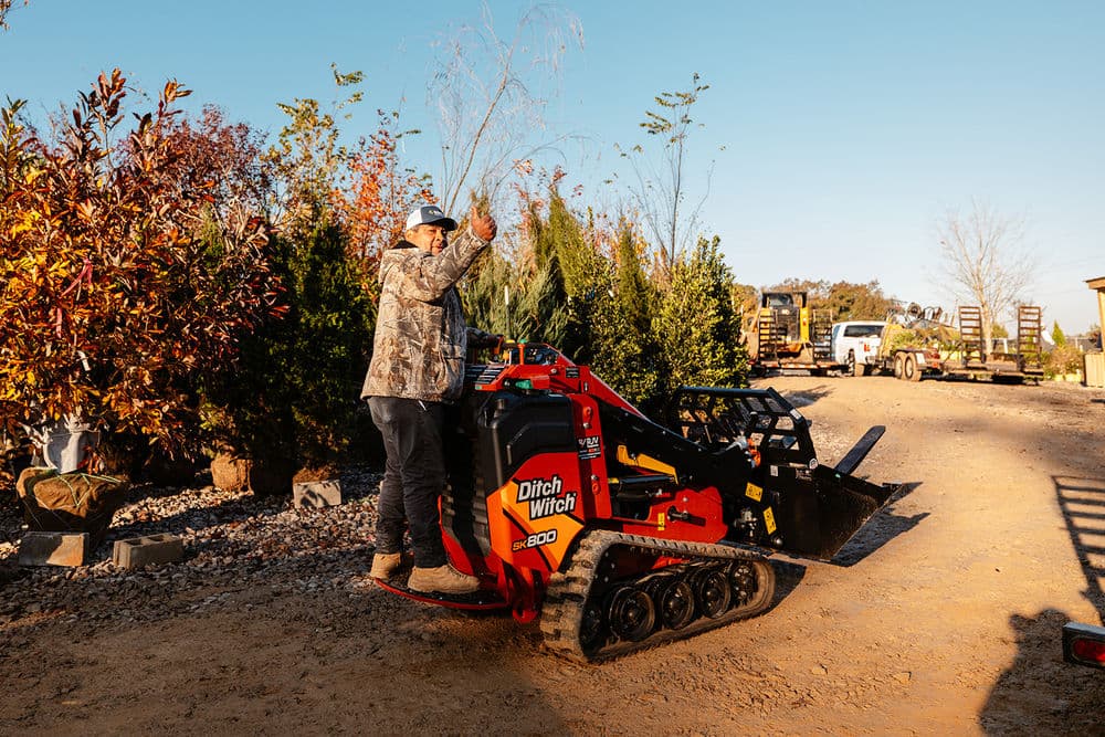 Operator using a Ditch Witch SK900 mini skid steer in a landscape nursery with trees.