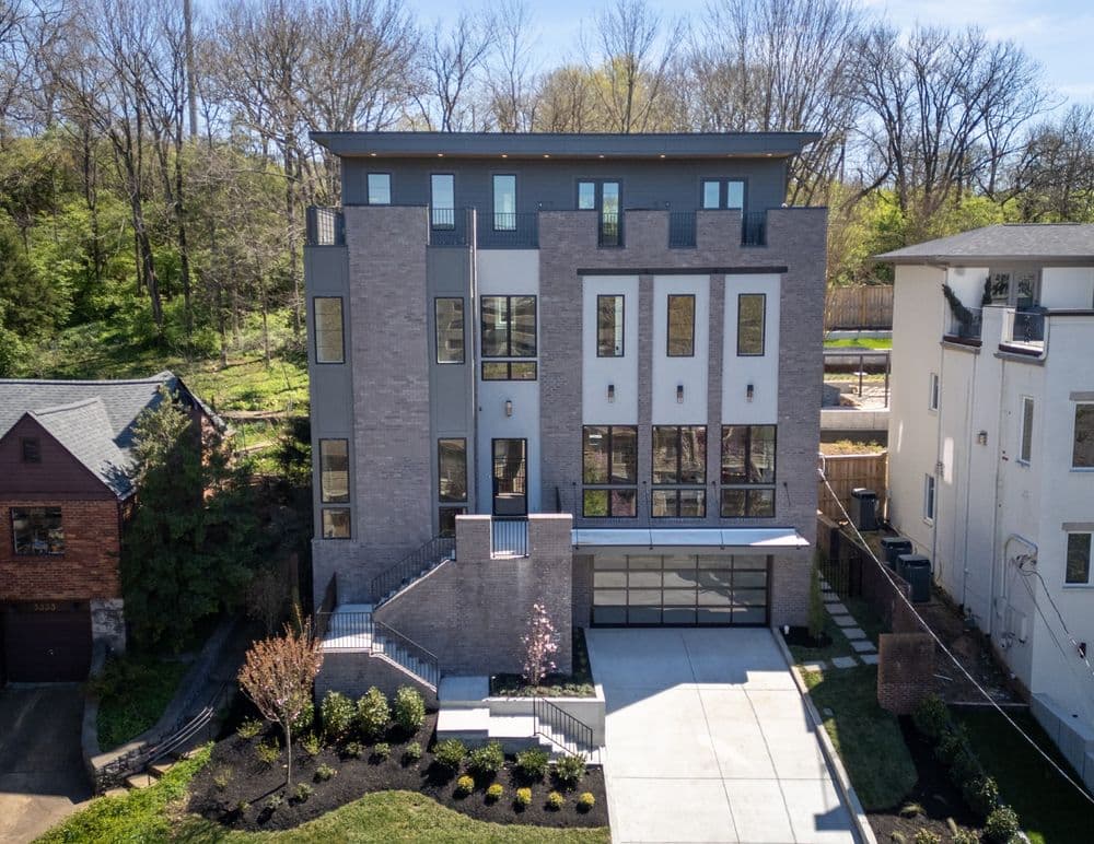 Modern three-story brick home with large windows, driveway, and landscaped garden.